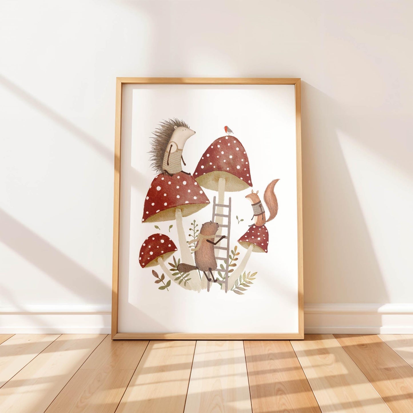 A single framed art print leaning against a white wall. The whimsical watercolor illustration features a hedgehog, squirrel, and beaver playing on large red and white mushrooms (toadstools).
