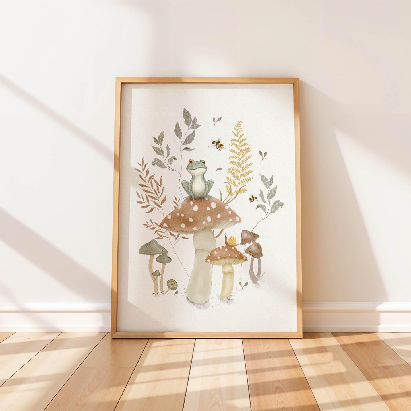 Frog on mushroom art print in a light wood frame on a wood floor, showing a frog sitting on a large mushroom with smaller mushrooms, ferns, and bees surrounding it