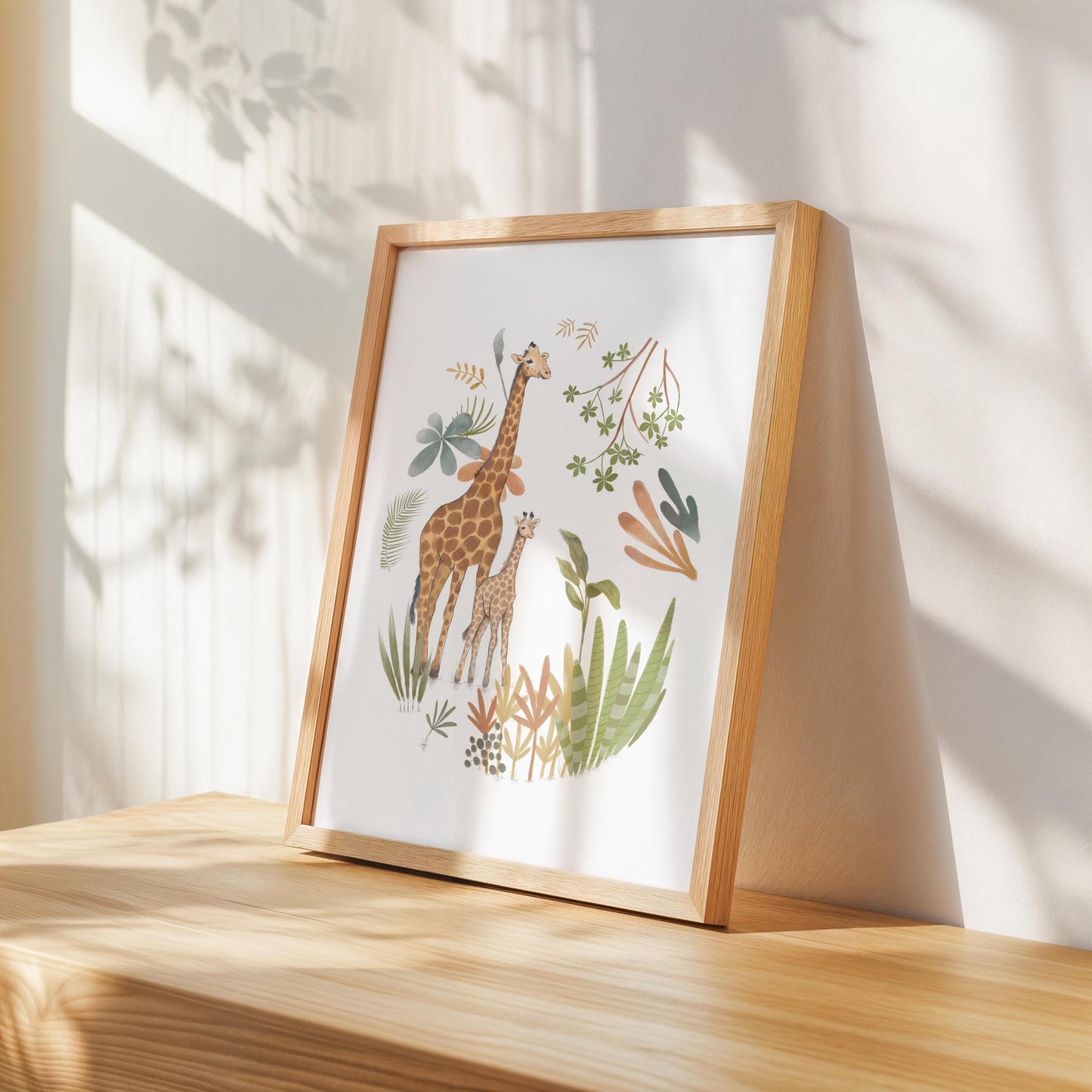 A framed illustration of two giraffes surrounded by various plants and foliage, placed on a wooden surface against a white wall.